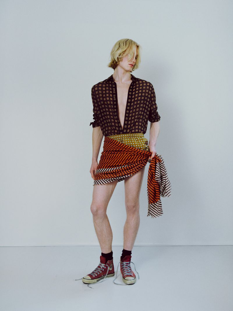 Young adult stands against a white wall wearing an unbuttoned patterned shirt, a red-striped wrap-skirt, and untied red sneakers.