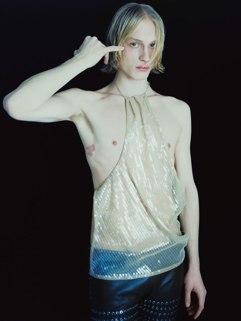 Androgynous fashion model with blond hair in sequin halter top and black studded shorts poses against a dark background.