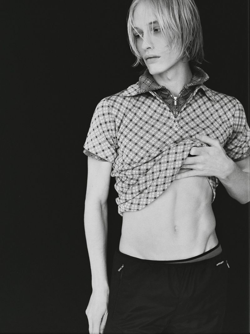Monochrome portrait of a slender adult man with a partially raised plaid shirt against a solid black background in eye-level medium shot.