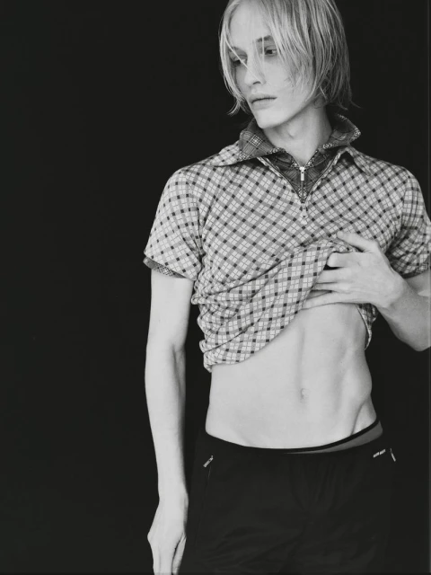 Monochrome portrait of a slender adult man with a partially raised plaid shirt against a solid black background in eye-level medium shot.
