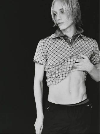 Monochrome portrait of a slender adult man with a partially raised plaid shirt against a solid black background in eye-level medium shot.