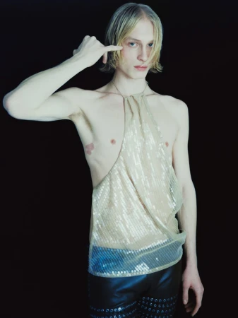 Blonde male model wearing a sheer sequin halter top and dark trousers against a plain black studio background.