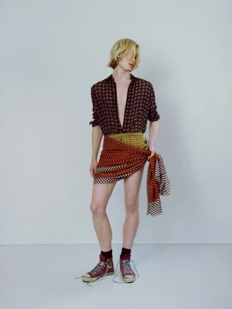 Blonde male model wearing patterned shirt and wrapped striped skirt against plain backdrop for Behind the Blinds