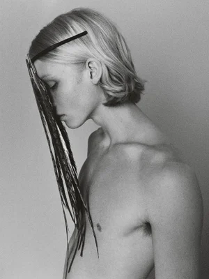 Black and white portrait of a shirtless male model with long blonde hair wearing a feathered headpiece covering his face.