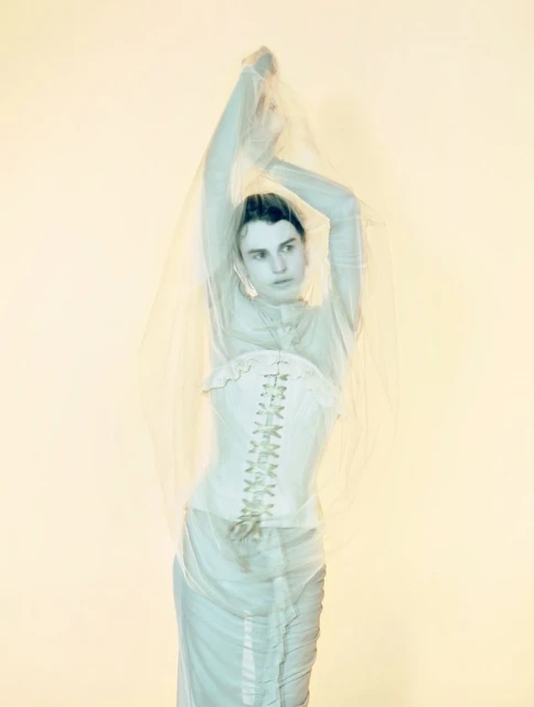 Full-length shot of a woman wearing a dress and corset, with her arms raised against a neutral background in a fashion studio.