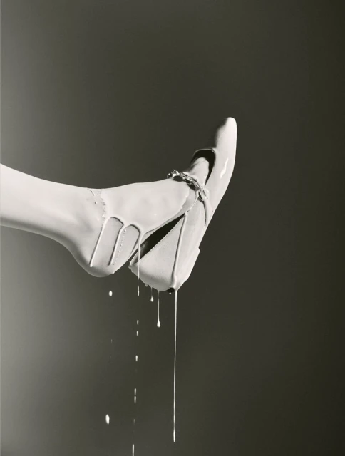 Elevated view of a high-heeled shoe covered in dripping white paint against a neutral gray background in a studio shot.
