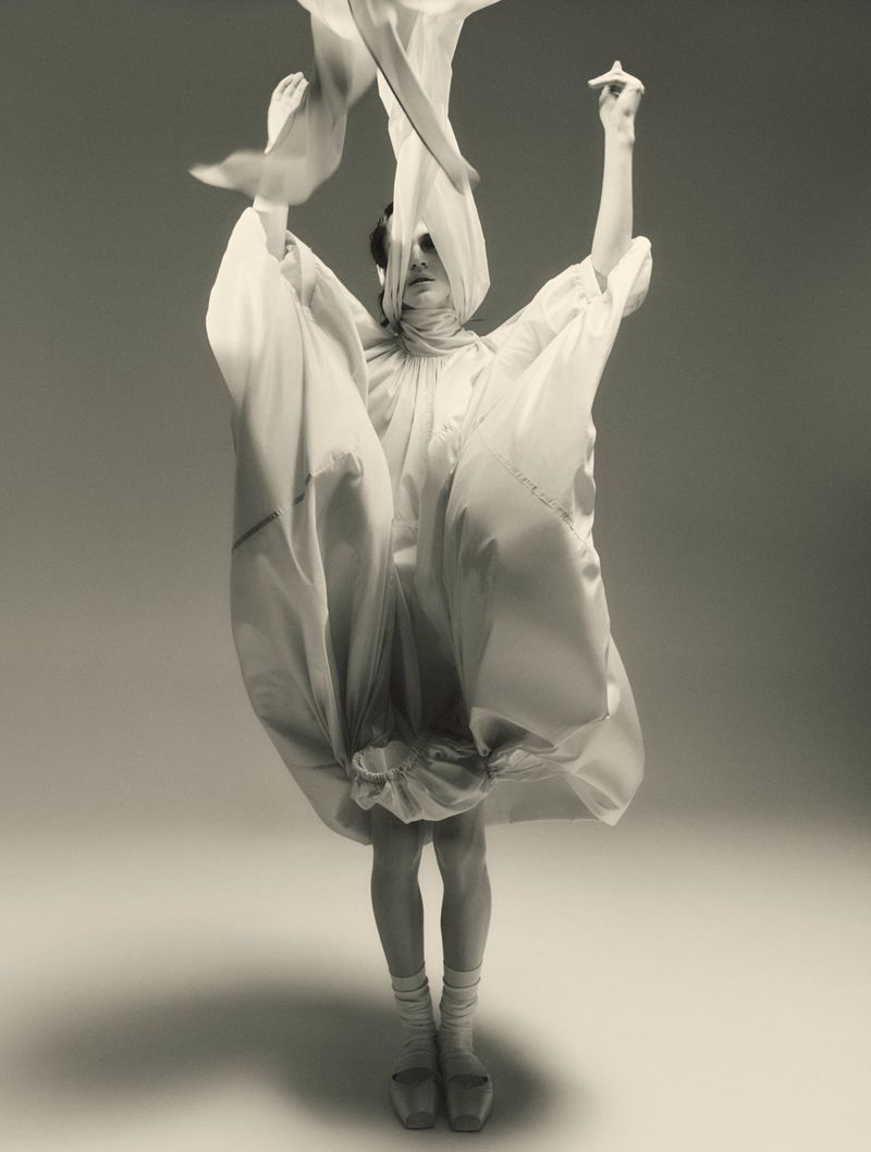 Model in voluminous white dress with floating fabric and head covering against a neutral seamless backdrop