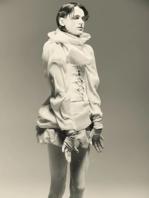 A person in an ornate garment stands in a studio, water dripping from their hands, creating an artistic and somewhat surreal visual.