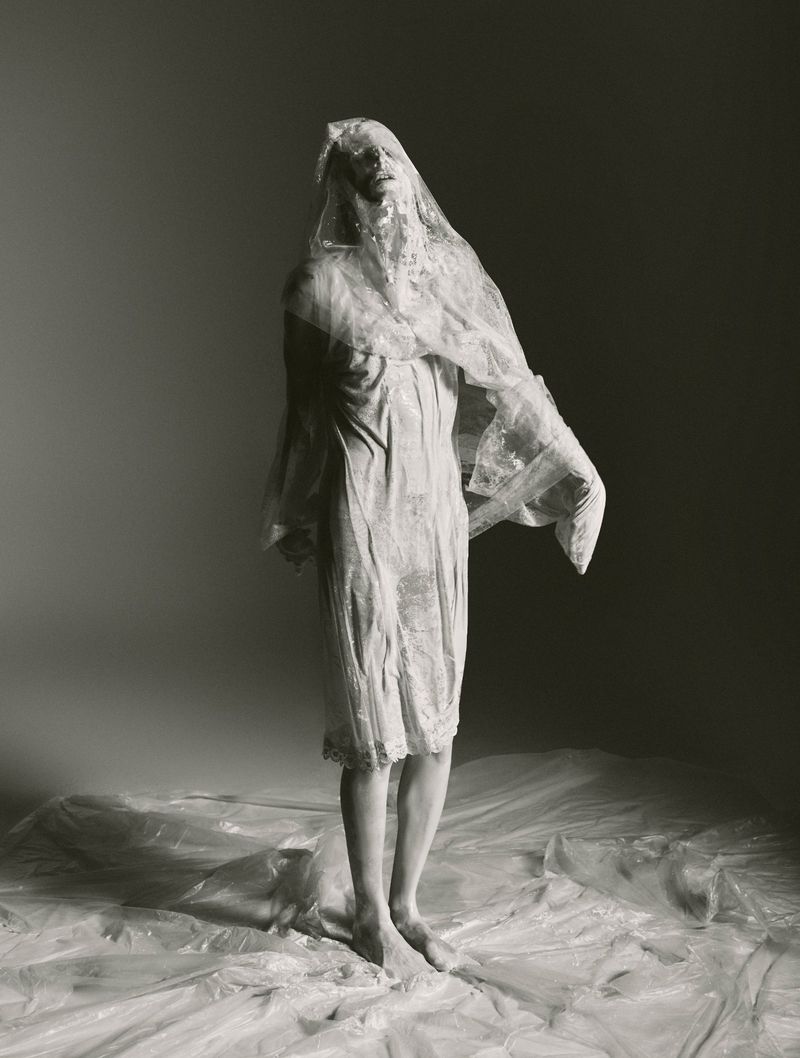 Full-length portrait of a person in a white dress and sheer covering, standing barefoot on plastic sheeting against a neutral backdrop.