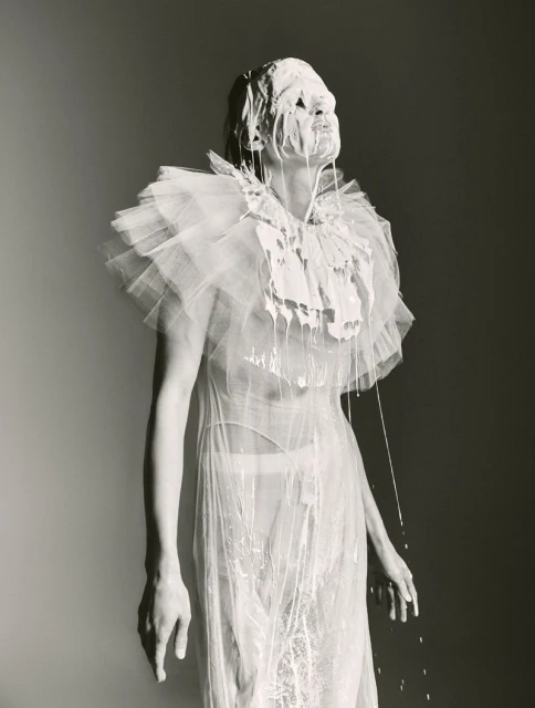 Artistic black and white portrait of an adult female covered in white fluid wearing a translucent ruffled collar and dress in a studio.