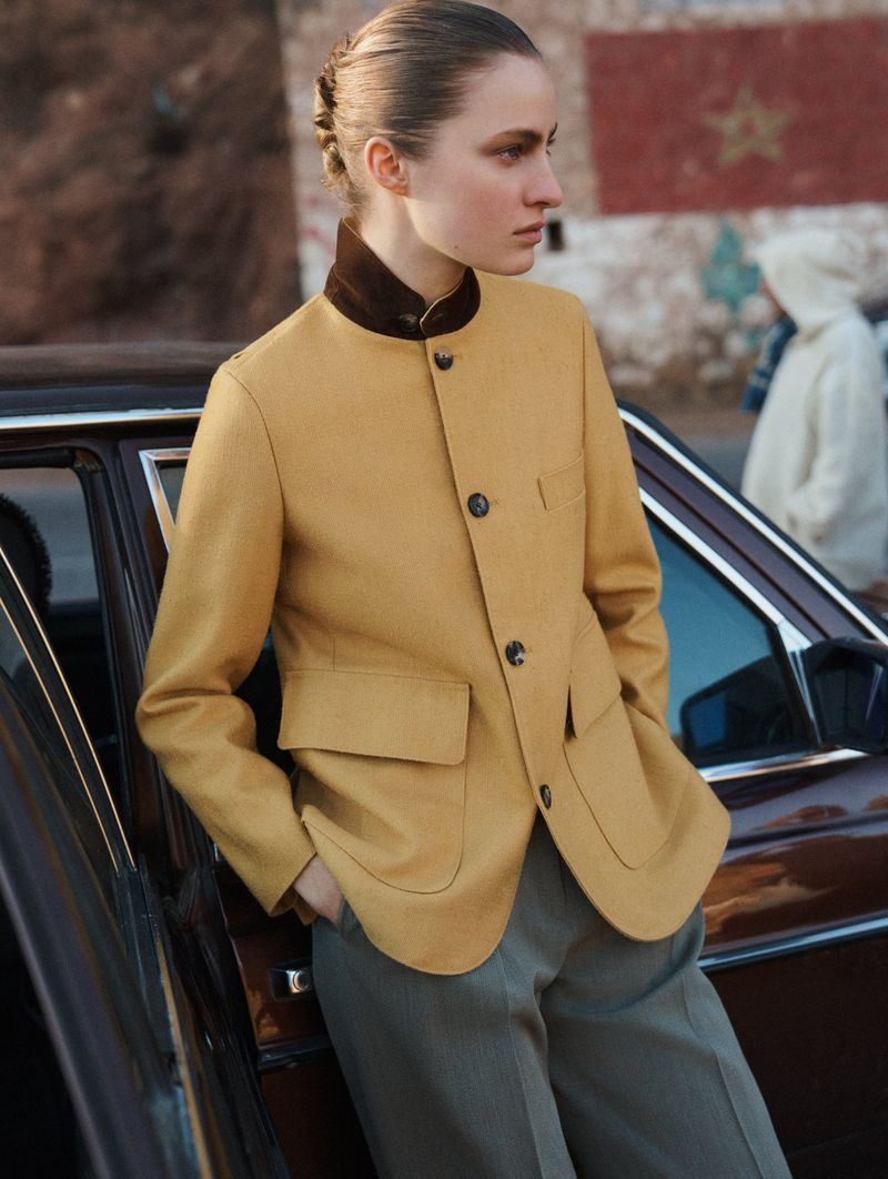 A fashionable woman in a mustard-colored blazer stands next to a vintage car with a brick wall and a person in the background.