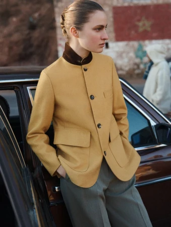 A fashionable woman in a mustard-colored blazer stands next to a vintage car with a brick wall and a person in the background.