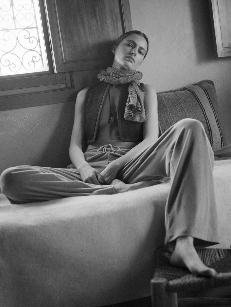 Monochrome portrait of a slender woman in lounge wear, sitting on a blanket-covered bed in an indoor setting with natural light.