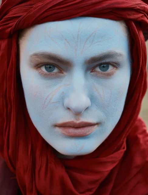 Close-up portrait of a woman with face paint and a red headscarf. Her blue eyes and painted face are the dominant features of the image.