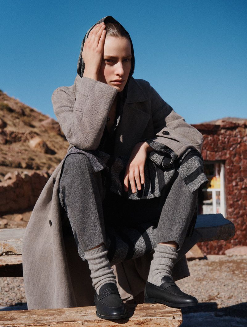 A woman in a grey coat and headscarf sits on a stone structure in front of a mountain and building in a fashion editorial shot.
