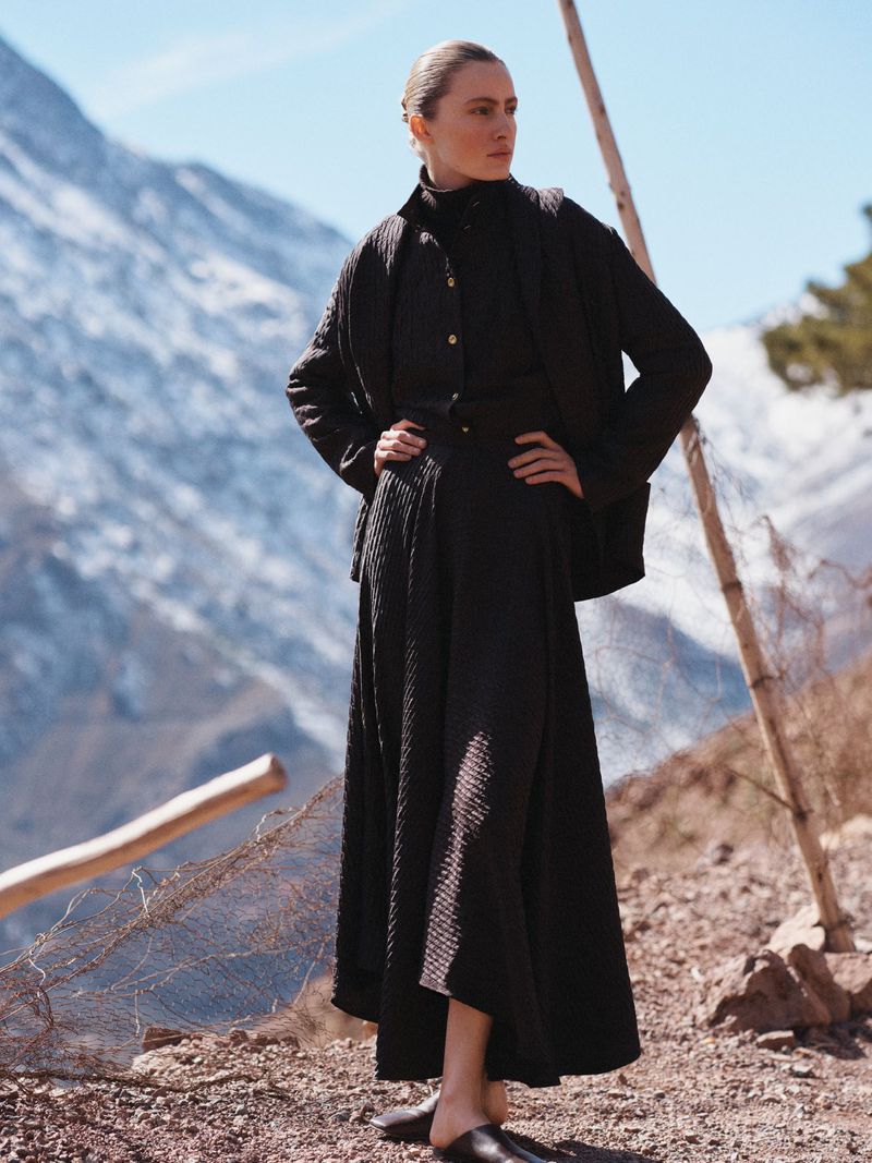 Model Felice Nova Noordhoff posing in dark textured clothing on a rocky mountain slope with snowy peaks behind