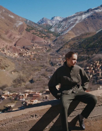 Woman poses in an olive-green outfit against a mountainous landscape backdrop, featuring clear blue skies and traditional village architecture.