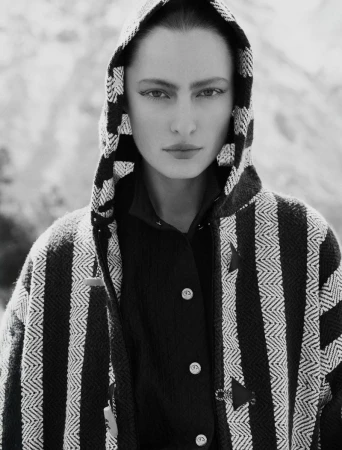 Black and white portrait of a woman with a hooded striped jacket against a blurred outdoor background, creating an editorial fashion image.