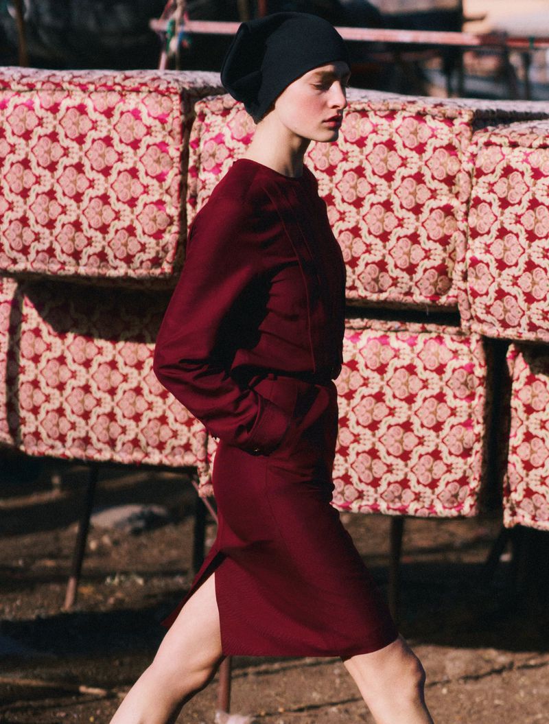 A woman in a long-sleeved burgundy dress and black cap is walking on a dirt path. Stacks of patterned cushions in the background.