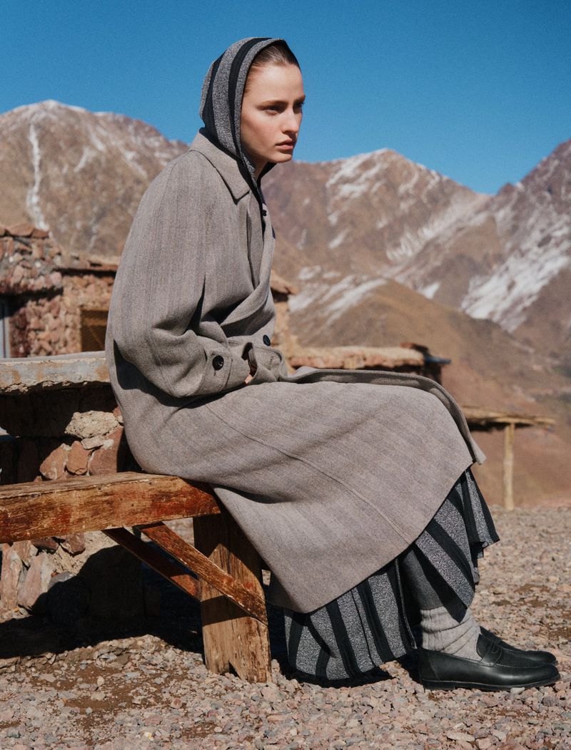 A woman sits on a weathered wooden bench in a full-length grey wool coat and striped headscarf, set against a desert mountain range.
