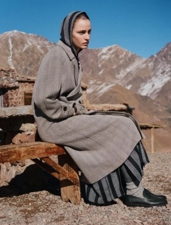 A woman sits on a weathered wooden bench in a full-length grey wool coat and striped headscarf, set against a desert mountain range.