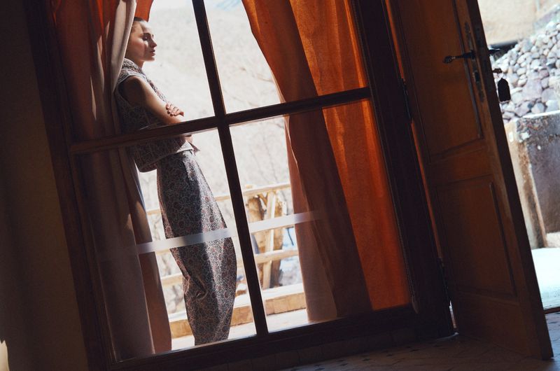 Adult female posing in a patterned two-piece outfit, leaning against a window with orange curtains in a sunlit, rustic interior.