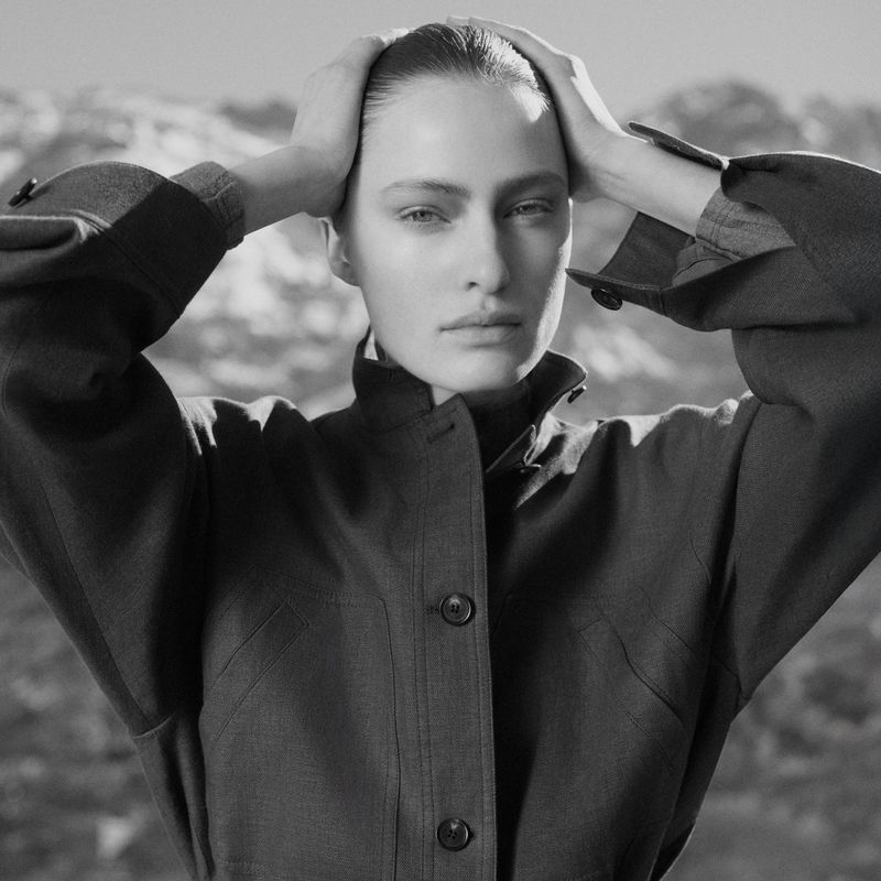 Black and white portrait of a woman posing in front of a mountain range, wearing a coat and with her hands on her head.