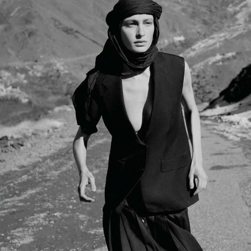 Monochrome fashion shot of a woman wearing a headscarf, vest, and skirt, posing on a road in a mountainous environment with soft lighting.