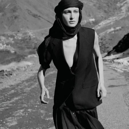 Monochrome fashion shot of a woman wearing a headscarf, vest, and skirt, posing on a road in a mountainous environment with soft lighting.
