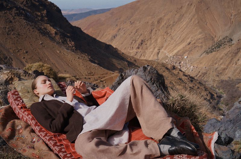 Woman rests on a blanket with patterned fabric in a mountainous landscape. She's wearing wide-leg pants and a dark brown blazer.