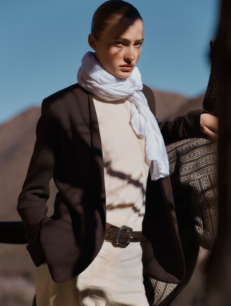 Fashion shot of a stylish woman in a blazer and scarf, posing against a backdrop of mountains under a clear sky, medium shot.