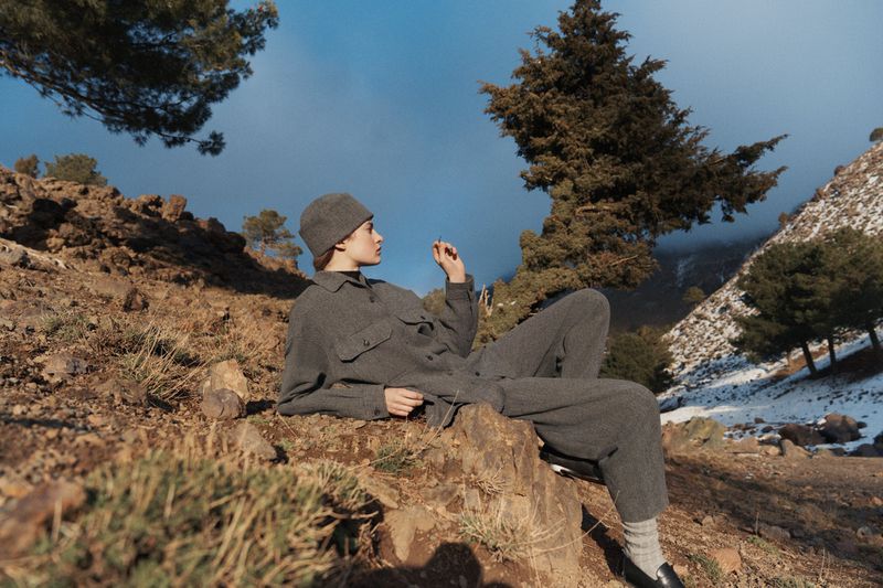 Woman in a gray wool suit and beanie lying on a rocky hillside with sparse vegetation in the mountains against a blue sky.