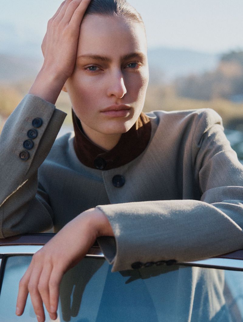 Portrait of an adult woman with a neutral facial expression, leaning on a car. She is wearing a jacket and collared shirt outdoors.