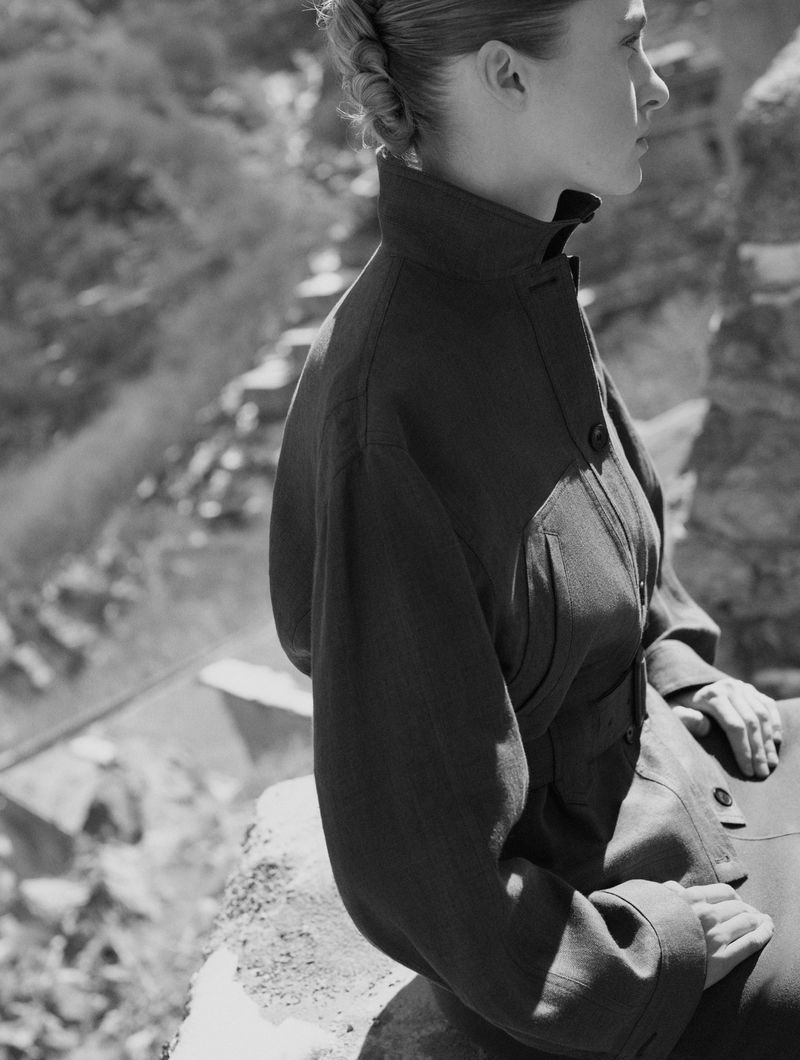Monochrome shot of a stylish adult female in a jacket, seated outdoors against a rugged, mountainous background, looking off to the side.