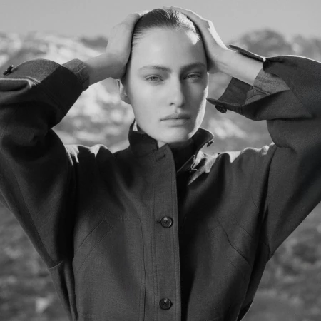 Close-up portrait of model Felice Nova Noordhoff holding her head with both hands, wearing a dark buttoned jacket against an out-of-focus background.