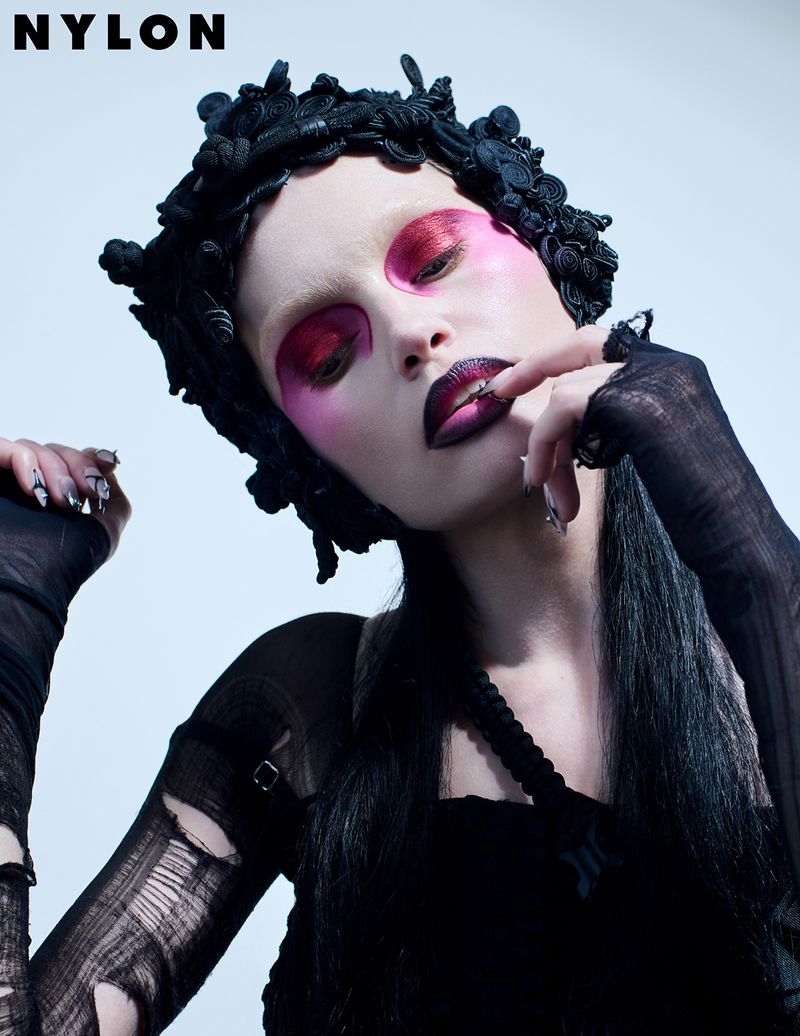 Dark and moody beauty shot features a woman with magenta eye makeup, dark lipstick, and a floral headpiece against a light background.