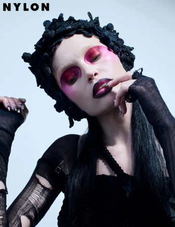 Dark and moody beauty shot features a woman with magenta eye makeup, dark lipstick, and a floral headpiece against a light background.