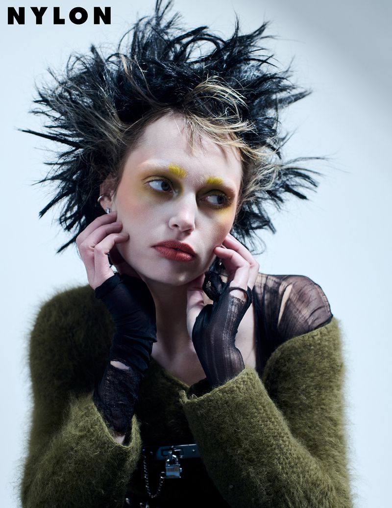 Editorial fashion shot of a woman with creative makeup and spiky hair wearing an olive sweater and black gloves, hands near face, against white background.