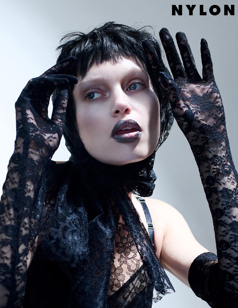 Fashion editorial shot of a model wearing black lace gloves and top, accessorized with a lace scarf and bold black lipstick, posing in a studio.