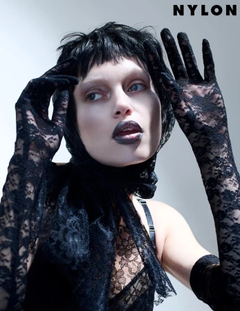 Fashion editorial shot of a model wearing black lace gloves and top, accessorized with a lace scarf and bold black lipstick, posing in a studio.