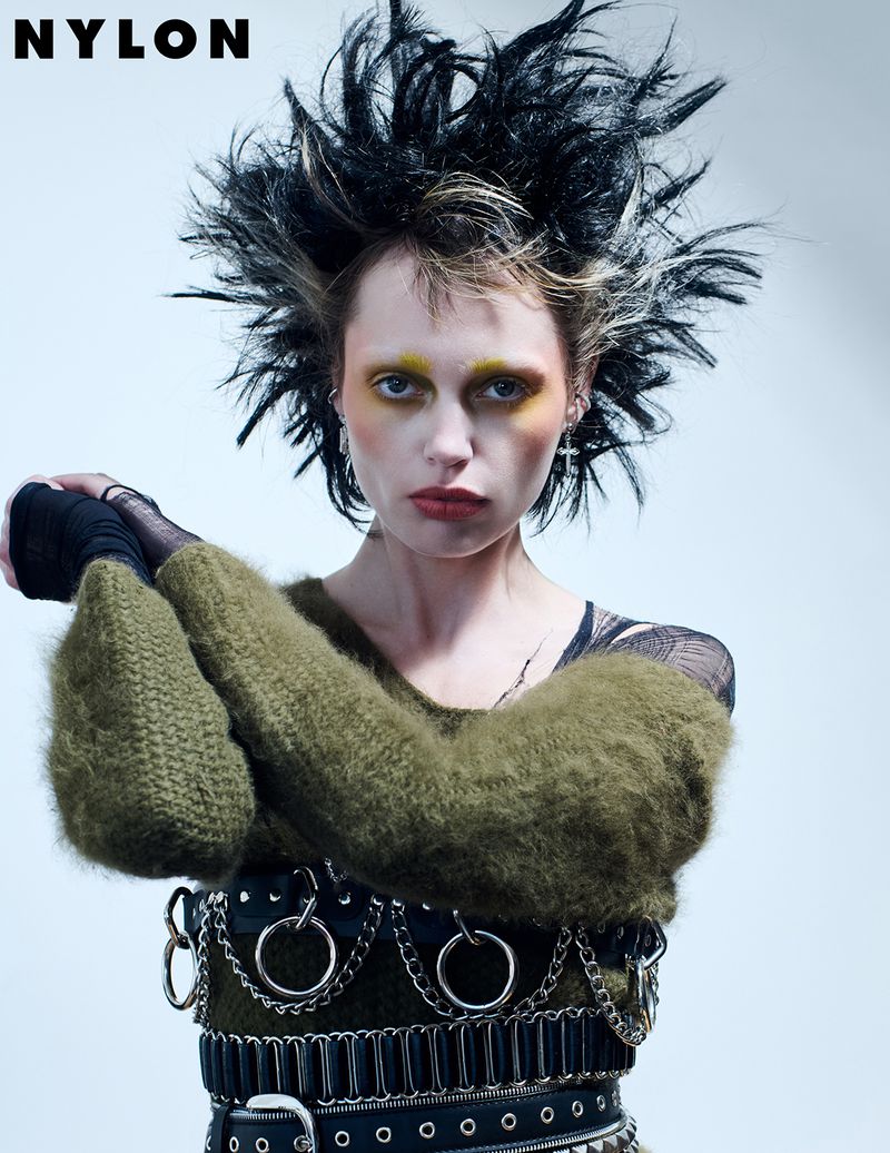 Editorial fashion shot of a female model with dramatic black and blonde feathered hair, heavy makeup, and a studded and chained belt.