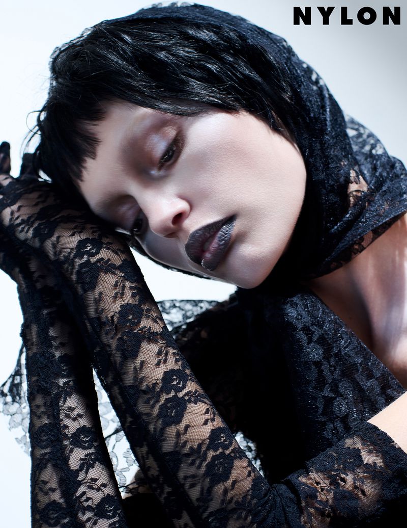 Portrait of a young woman with dark makeup and lace, wearing a black lace head covering, against a light background, by NYLON Magazine.