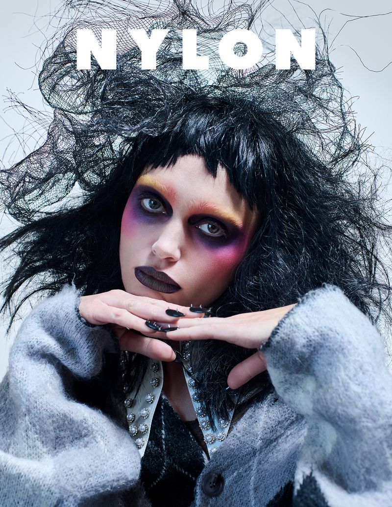 Portrait of a woman with creative dark makeup and black wig, wearing a grey cardigan, with the magazine title 'NYLON' displayed above.