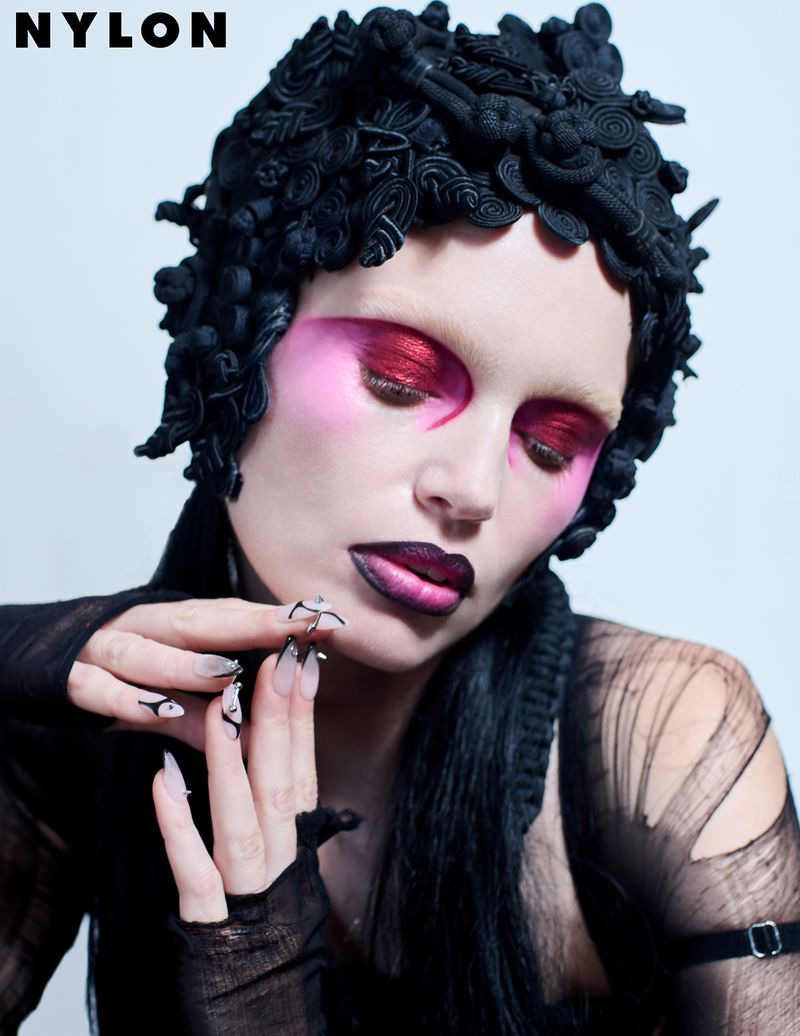 Close-up portrait of a woman with dramatic makeup, a black headpiece, and long nails, posing against a white backdrop, as seen in Nylon.