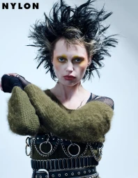 Model Staz Lindes with spiky black hair, yellow eye makeup, and green sweater against a white backdrop for Nylon Germany