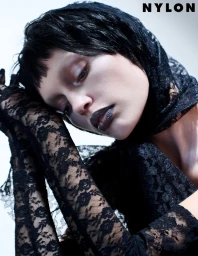 Model Staz Lindes wearing a black lace headscarf and matching long lace gloves against a white studio background.