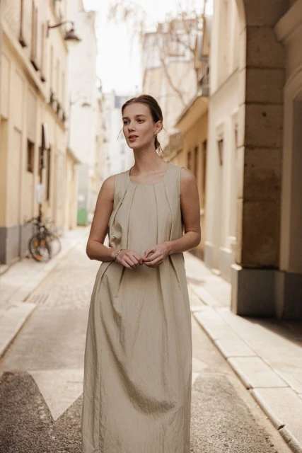 A woman in a long beige dress stands in a narrow alleyway with buildings on both sides and a bicycle in the background.