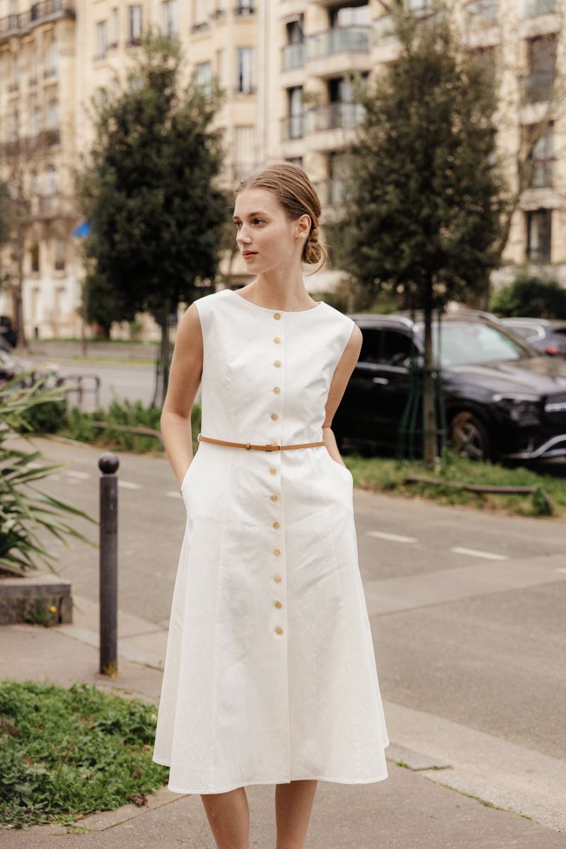 Woman in white midi dress with brown belt stands on city sidewalk with apartment buildings and parked cars in the background.