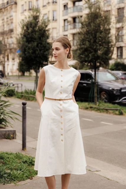Woman in white midi dress with brown belt stands on city sidewalk with apartment buildings and parked cars in the background.