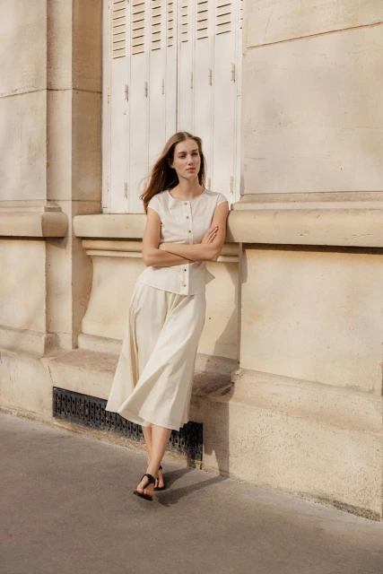 A woman stands outside a building with cream-colored walls and white shutters, wearing a matching cream-colored top and pants.